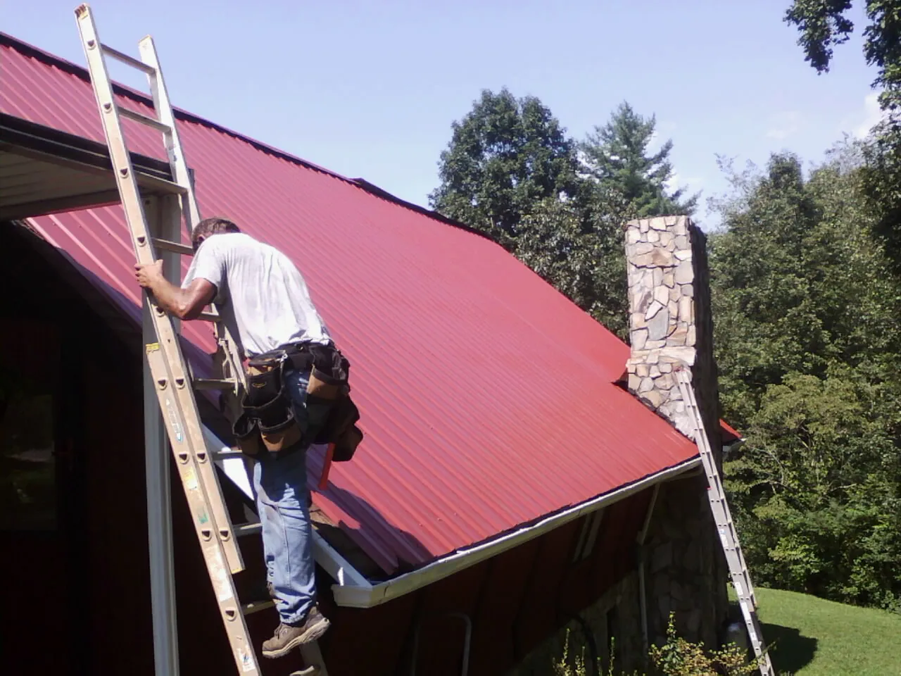 Roofer on a ladder beside a red metal roof and stone chimney — Soffit & Fascia Repair by Roofing Brothers in Ruby